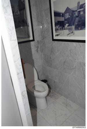 This is an evidence photograph showing the interior of a bathroom featuring marble tile walls, a white toilet, and framed architectural photographs on the wall. The image appears to be documentation of a property interior, likely from one of Epstein'