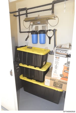 This photograph documents a storage closet or utility room containing stacked black and yellow storage containers, water filtration equipment including blue filter cartridges, black piping infrastructure, and various tools and instructional materials