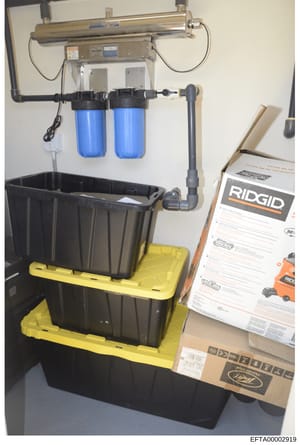 This photograph documents a water filtration system installed on a wall, featuring dual blue filter cartridges, UV sterilization unit, and collection containers. The image includes RIDGID brand documentation and cardboard packaging visible in the fra