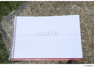 This photograph shows a spiral-bound notebook page with the word 'gazebo' written in blue pen. The notebook is positioned on what appears to be a concrete or stone surface outdoors. The minimal text and casual nature of the document suggest it may be