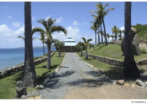 This photograph shows a seaside residential property featuring a distinctive white dome-roofed building, manicured grounds, and a palm tree-lined gravel driveway leading to the main structure. The property sits on elevated oceanfront land with stone 