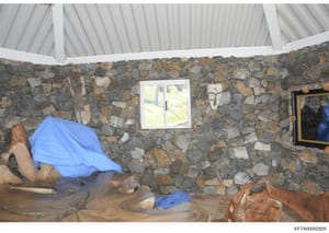 This is an evidence photograph showing the interior of a room with distinctive rough stone walls, a window, and furniture covered with blue tarps. The image appears to be from a property inspection or documentation, possibly related to one of Epstein