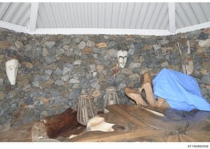 This is a photograph of an interior space featuring a rustic stone wall with decorative African masks mounted as wall art, wooden sculptures, woven baskets, and other artifacts displayed on wooden furnishings. The image appears to be from one of Jeff