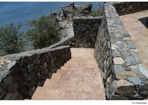 This photograph shows a waterfront property featuring a terracotta tile walkway bordered by natural stone masonry walls, with Mediterranean-style construction overlooking blue water. The image appears to be documentation of property infrastructure, l