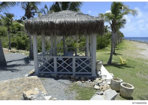 This photograph shows a decorative gazebo with thatched palm roof located on beachfront grounds, surrounded by palm trees and manicured lawn. The structure features wooden lattice panels and overlooks a coastal shoreline. The image appears to be evid