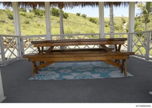 This is a photograph of an interior patio or covered gazebo area featuring a wooden bench positioned on a decorative blue and white patterned rug. The space overlooks lush, green hillside terrain with palm trees visible through screened windows and d