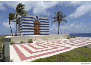 This is a photograph of a unique oceanfront property featuring a striking modernist building with blue and white horizontal stripes and a large geometric wooden entrance door. The property includes manicured grounds with red and white geometric pavin