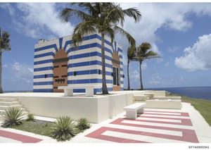 This is a photograph of a distinctive modern building with bold blue and white horizontal stripes and a stylized brown central design element, located on an oceanfront property. The structure features palm trees, manicured landscaping, and a striped 
