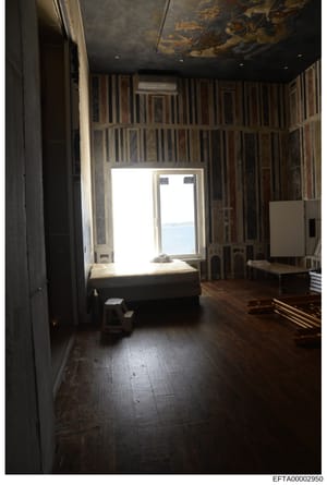 This is a photograph of an interior room, likely a bedroom or living space, showing ornate striped wall paneling, a dark ceiling with decorative details, wooden flooring, and large windows offering a view of water. The room appears to be part of a re