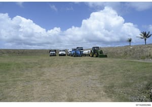 This photograph shows a rural property or ranch with multiple vehicles and agricultural equipment lined up in an open field. The image depicts what appears to be a working ranch or agricultural facility with tractors, trucks, and other heavy equipmen