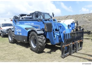 This is a photograph of a blue Genie GTH-1256 telescopic handler (telehandler) heavy equipment vehicle, taken at an outdoor location with construction/industrial equipment visible in the background. The image documents equipment that may have been us