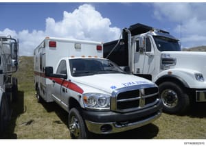 This photograph shows three emergency medical service (EMS) vehicles parked together in an outdoor, grassy area with hills in the background. The image displays a white ambulance with red and blue markings in the foreground, a white ambulance unit in