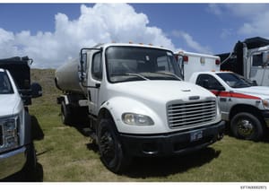 This is a photograph of a white Freightliner commercial tanker truck parked at what appears to be an equipment yard or fleet facility. The truck is photographed outdoors in daylight conditions alongside other commercial vehicles. The image may be par