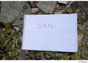 This photograph shows a spiral-bound notebook placed on pavement/ground with green vegetation visible around it. The notebook has 'GYM' handwritten in pen on its lined page. The document appears to be evidence photography, likely showing an item seiz