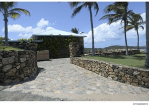 This is a photograph of a beachfront property featuring a stone structure with a metal roof, surrounded by palm trees and stone walls. The image shows a tropical or subtropical coastal location with clear views of water and vegetation in the backgrou