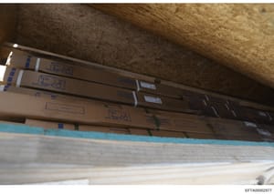 This is a photograph of a storage or construction area showing multiple cardboard boxes and rolled materials stacked against walls with concrete ceilings and wooden/paneled surfaces. The boxes bear labeling and shipping information, suggesting invent