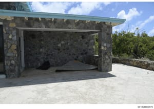 This is a photograph of an exterior structure featuring stone construction with a turquoise metal roof overhang. The building appears to be a pavilion or outbuilding with an open front design, situated in a tropical or subtropical location with veget