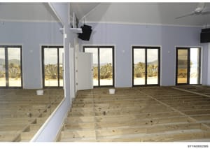 This is a photograph of an interior space under construction or renovation, featuring a spacious room with perimeter glass doors and windows overlooking a desert landscape with palm trees and mountains. The room has light purple/lavender walls, woode