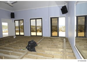 This photograph documents the interior of a beachfront residential property during active construction or renovation. The image shows a large open room with exposed wooden subfloor framing, light blue walls, black-framed glass doors and windows openi