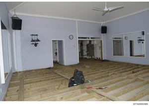 This photograph documents an interior room during renovation or construction, showing exposed wooden floor joists, freshly painted light blue walls, ceiling fans, and various interior fixtures. The image appears to be evidence documentation from a pr
