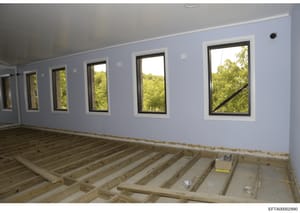 This is a photograph of an interior room under construction or renovation, featuring light blue-painted walls, multiple large black-framed windows overlooking wooded landscape, and exposed wooden floor joists. The image appears to be documentation of