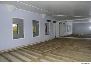 This photograph shows an interior space under construction or renovation, featuring unfinished wooden flooring, light blue/lavender painted walls, and multiple framed openings or doorways. The space appears to be a large open area with recessed light