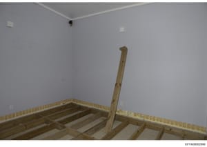 This photograph shows an unfinished residential room during construction, featuring exposed wooden floor joists, framing studs, and white drywall walls. The image appears to be documentary evidence of property conditions or renovation work, likely ta