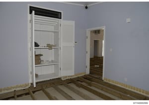 This is a photograph of an interior room in an early stage of renovation or construction. The space features exposed wooden floor joists, light blue/periwinkle walls, white trim and doors, and built-in shelving with stored items. The image appears to