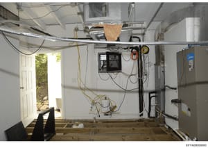 This is a photograph of an unfinished basement room showing electrical panels, HVAC equipment, exposed wiring, and a doorway with outdoor visibility. The image appears to be documentation of a property's mechanical systems and infrastructure, likely 