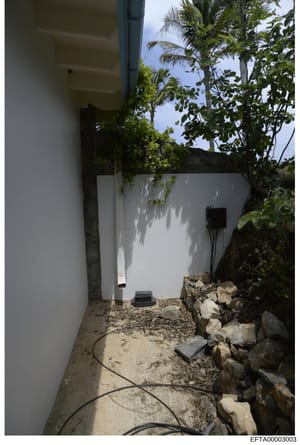 This is a photograph of a narrow exterior passage or walkway at a property, showing white-painted walls, landscaping with tropical vegetation (palm trees visible), and scattered rocks along the right side. The image appears to be evidence documentati