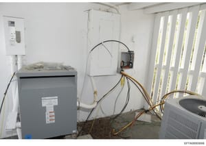 This photograph shows an HVAC system installation in progress within a property, featuring a gray transformer/electrical box on the left, exposed wiring, electrical panels, and air conditioning units. The image documents the mechanical and electrical