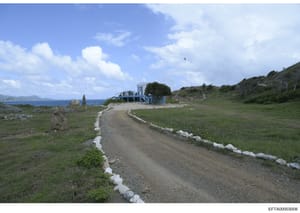 This photograph shows a remote coastal property with a distinctive blue and white building situated on elevated terrain overlooking the ocean. A dirt driveway with white stone borders leads to the structure, which appears to be located on an island o