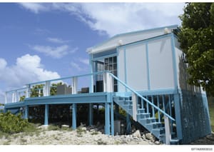 This is a photograph of a modern beachfront residential structure featuring blue and white exterior paneling, elevated on stilts above sandy terrain. The building appears to be a contemporary beach house or property with an open-air deck, multiple le