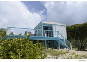 This photograph shows a modern elevated residential structure painted in light blue and white, situated on a beachfront property. The house features a raised foundation typical of coastal construction, large windows, and a wraparound deck or balcony.
