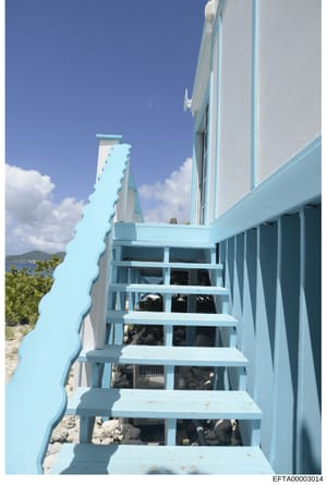 This is an evidence photograph showing the exterior of a modern coastal property with distinctive turquoise and white trim. The image captures an open staircase leading upward along the side of a building, with an ocean or water view visible in the b