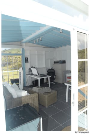 This is a photograph of a contemporary home office or studio space featuring a distinctive blue beam ceiling, work desk with computer equipment, comfortable seating area with woven furniture, and large windows overlooking natural landscape. The space