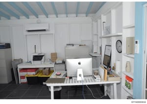 This is a photograph of an office or administrative workspace, featuring a white desk with an Apple computer, printer, filing cabinets, and various office supplies. The room has blue-and-white striped ceiling beams, white walls, dark flooring, and wa