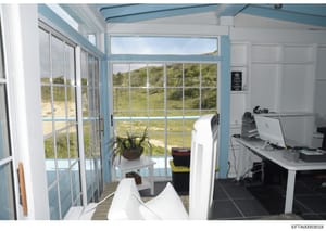 This photograph shows an interior room with extensive windows overlooking a coastal landscape with green hills and water visible in the background. The room appears to be a modern office or study space with computer equipment, an air conditioning uni