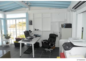 This is a photograph of a modern office workspace featuring a white desk, black office chair, computer equipment, storage shelving, and large windows overlooking a coastal landscape. The room appears to be part of a property investigation file, likel