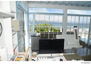 This photograph shows an interior office or workspace in a beachfront property, featuring floor-to-ceiling windows with panoramic ocean views, modern furnishings, and coastal landscape visible beyond. The image appears to be documentation of a reside