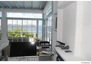 This is a photograph of a modern office/workspace interior with expansive waterfront views. The room features floor-to-ceiling glass doors and windows overlooking a bay or ocean with mountains visible in the distance, suggesting a coastal luxury prop
