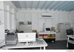 This is a photograph of a modern office space featuring white walls with blue ceiling beams, computer workstations, filing cabinets, and storage units. The room contains an HP laptop, printer, office supplies, and organizational materials in boxes an