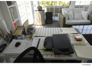 This photograph shows a bright, modern office space featuring a large work desk with computer equipment, office supplies, and documents. The room includes floor-to-ceiling windows with views to landscaping, wicker seating with white cushions, and var