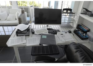 This is a photograph of a bright, modern residential office space containing a desk with scattered documents, a desktop computer setup, and office supplies. The room features large windows, contemporary furnishings including a white sofa with cushion