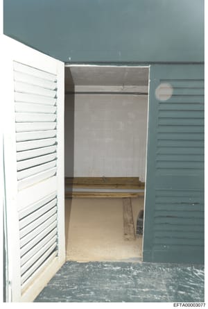 This photograph shows the interior of a small, empty room or closet space with teal-colored walls, white and teal louvered doors on either side, and what appears to be wooden shelving or structural elements inside. The room is bare and unfinished, ap