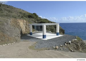 This is a photograph of a modern white structure with blue trim situated on a rocky beach overlooking the ocean. The building appears to be a small pavilion or shelter with a pergola-style roof, louvered windows, and concrete foundation, built into a