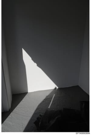 This is a black and white photograph showing an interior corner or architectural space with dramatic natural light streaming through an opening, creating geometric shadows on white and textured walls. The image appears to be a documentary or evidence