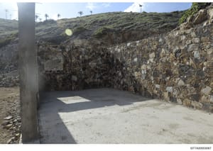 This photograph shows an outdoor stone structure with concrete pillars and walls situated on a hillside with arid vegetation and palm trees visible in the background. The structure appears to be under construction or renovation, with exposed stonewor