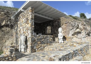 This photograph shows a stone building or structure built into a hillside, featuring two white sculptural figures positioned within and near the structure. The building appears to be constructed from stacked stone with a modern metal roof canopy. The