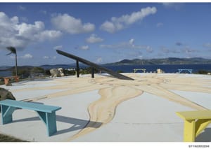 This photograph shows an expansive modern deck or terrace overlooking a blue ocean and mountainous coastline. The space features contemporary architectural elements including a large dark angular structure, colorful benches (turquoise and yellow), de